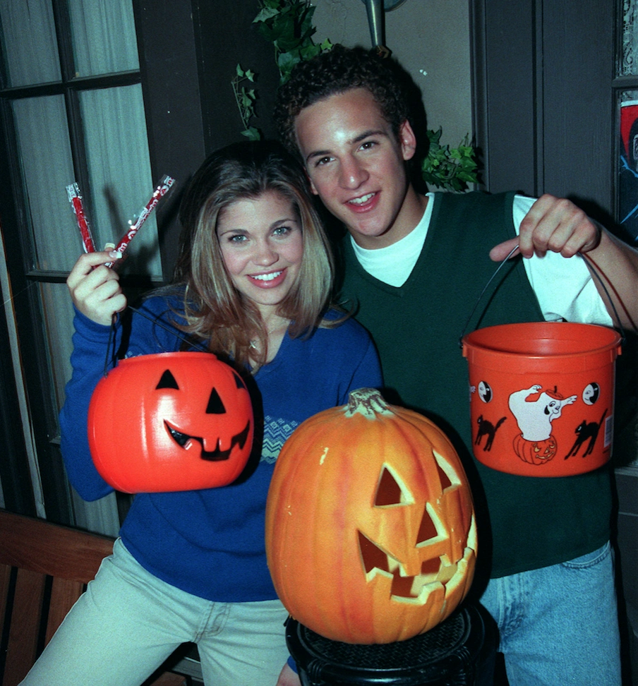 A young couple in casual 90s attire smiles as they hold bright orange pumpkin-shaped buckets filled with Halloween treats. They are standing in front of a carved jack-o'-lantern, capturing the nostalgic, fun spirit of Halloween during the 90s.