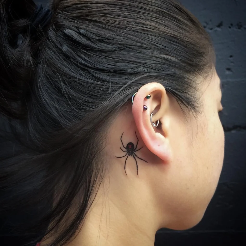 Side view of a realistic black widow spider tattoo behind the ear, paired with multiple ear piercings.