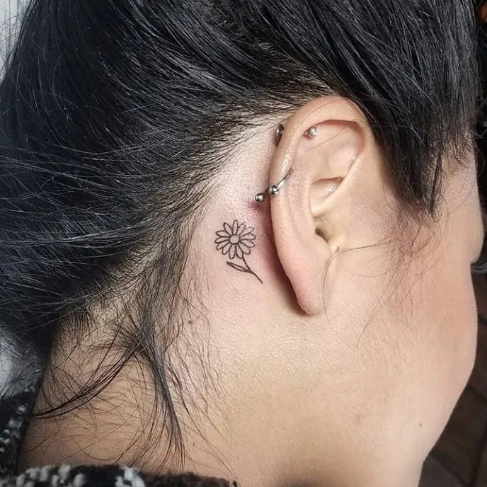 Side view of a small daisy tattoo behind the ear, paired with silver ear piercings.