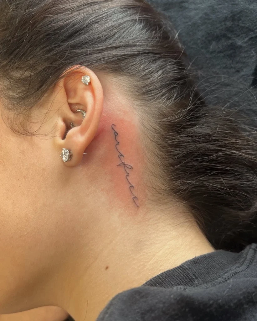 Side view of a minimalist "carefree" script tattoo behind the ear, accompanied by small ear piercings.