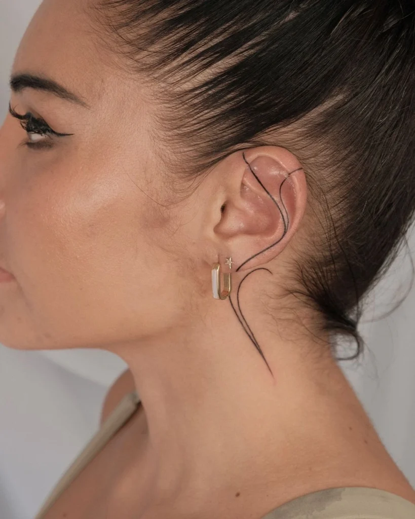 Side profile of a woman with a minimalist flowing line tattoo around the ear and extending to the neck, paired with small gold earrings.