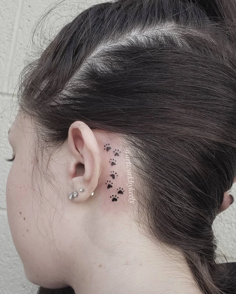 Side view of a paw print tattoo trail behind the ear, paired with small diamond stud earrings.