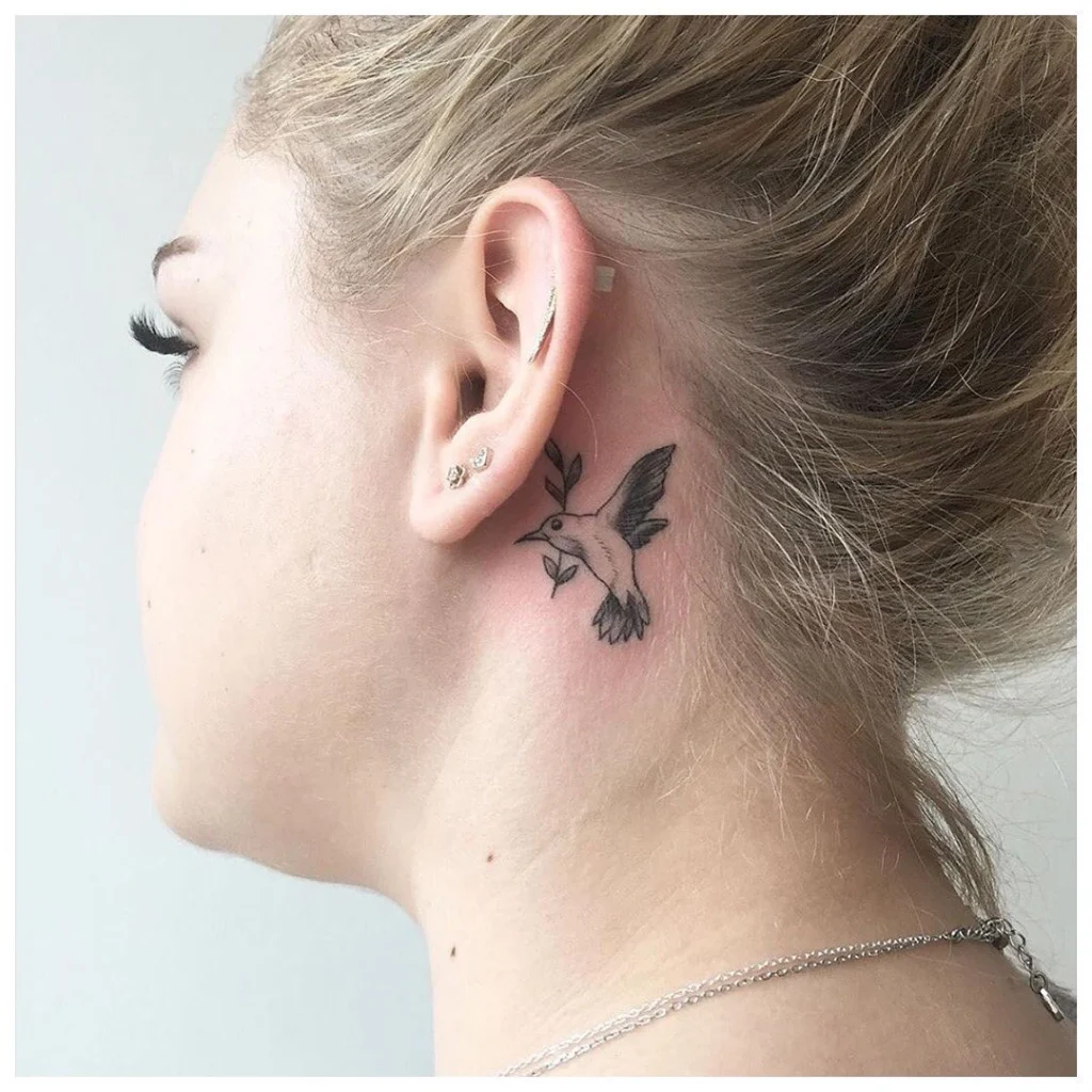 Side view of a hummingbird tattoo behind the ear, holding a branch, paired with small ear piercings.