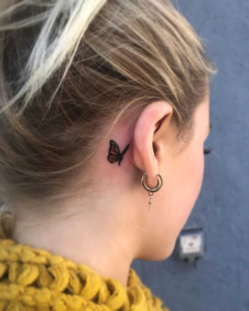 Side view of a small butterfly tattoo behind the ear, paired with a gold hoop earring.