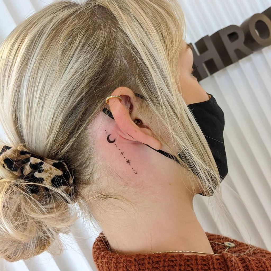 Side view of a celestial-themed tattoo with a crescent moon and stars behind the ear, paired with a small gold hoop earring.