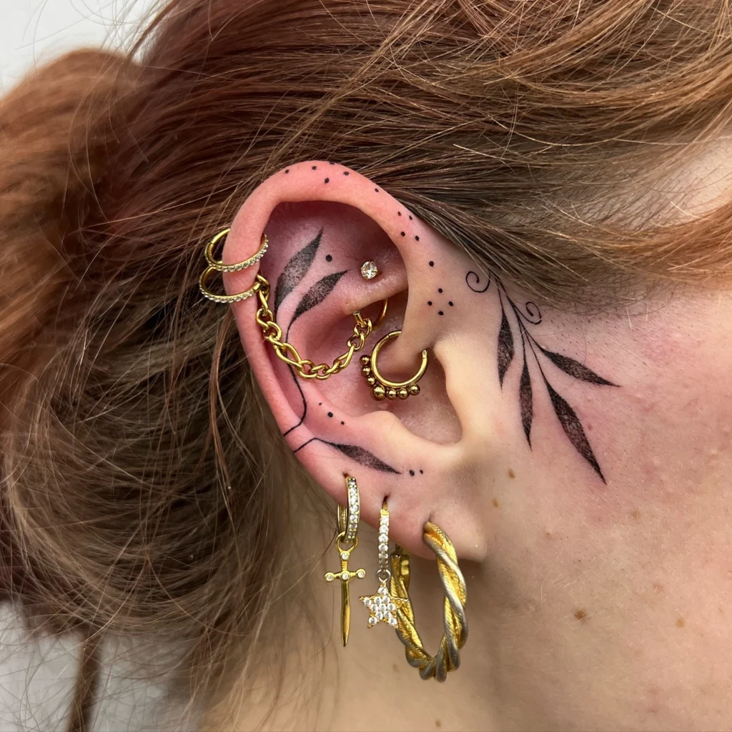 Close-up of an ear with a delicate leaf tattoo and multiple gold piercings, including hoops, chains, and charms.