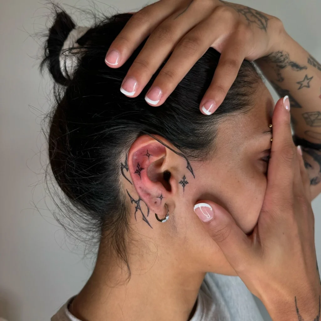 Side view of a person with a minimalist tattoo of vines and stars wrapping around the ear and extending to the neck and face, with a small hoop earring.