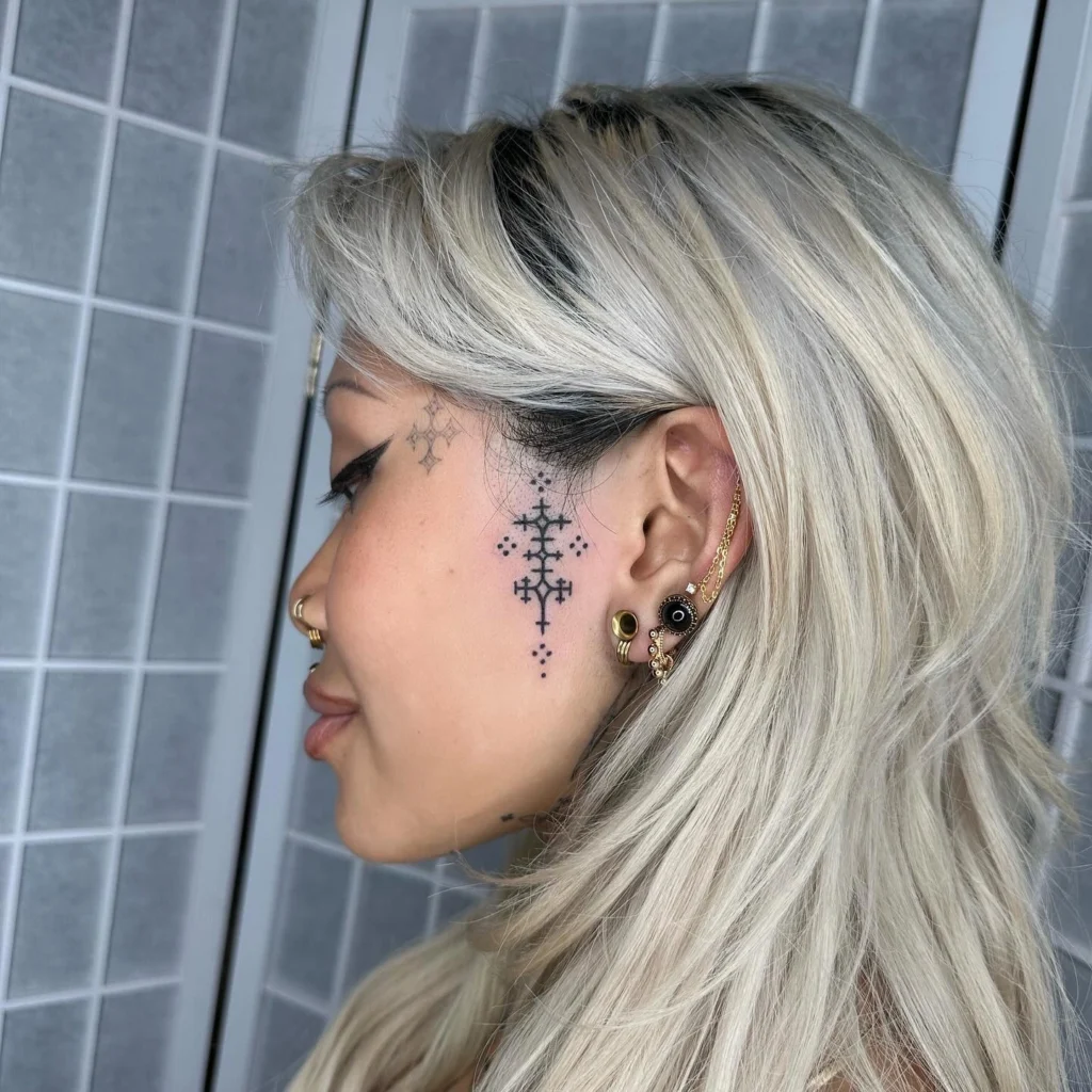 Side profile of a woman with a geometric and dotwork tattoo extending from her temple to the jawline, paired with gold ear piercings and blonde hair.