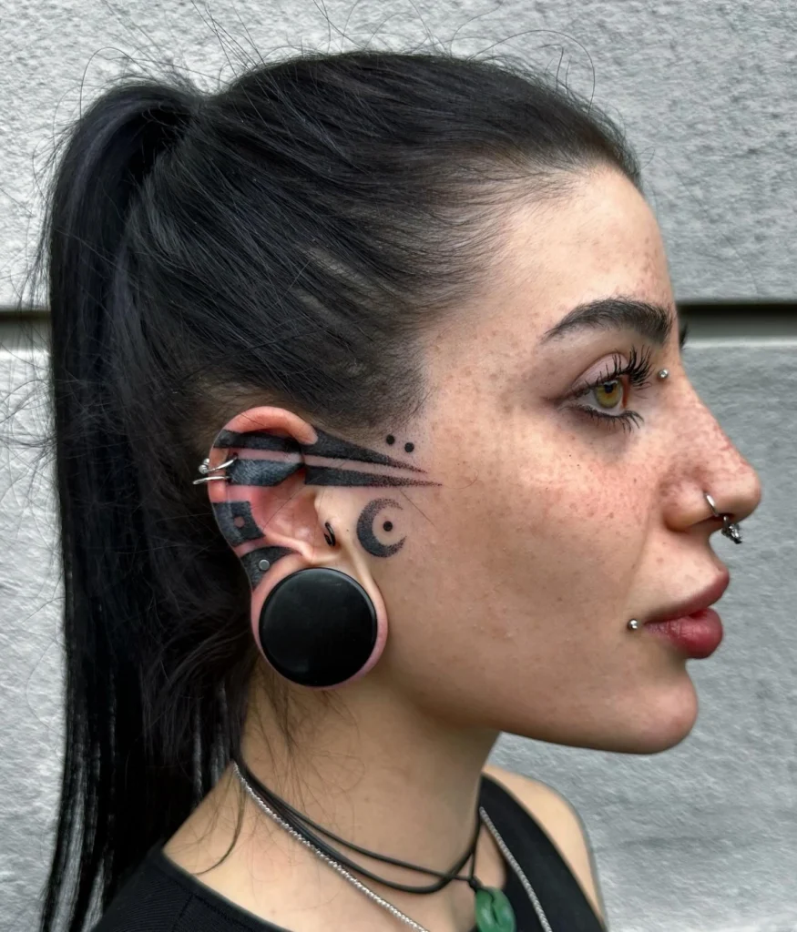 Side profile of a person with a bold geometric tattoo extending from the ear to the face, paired with large ear gauge and multiple facial piercings.