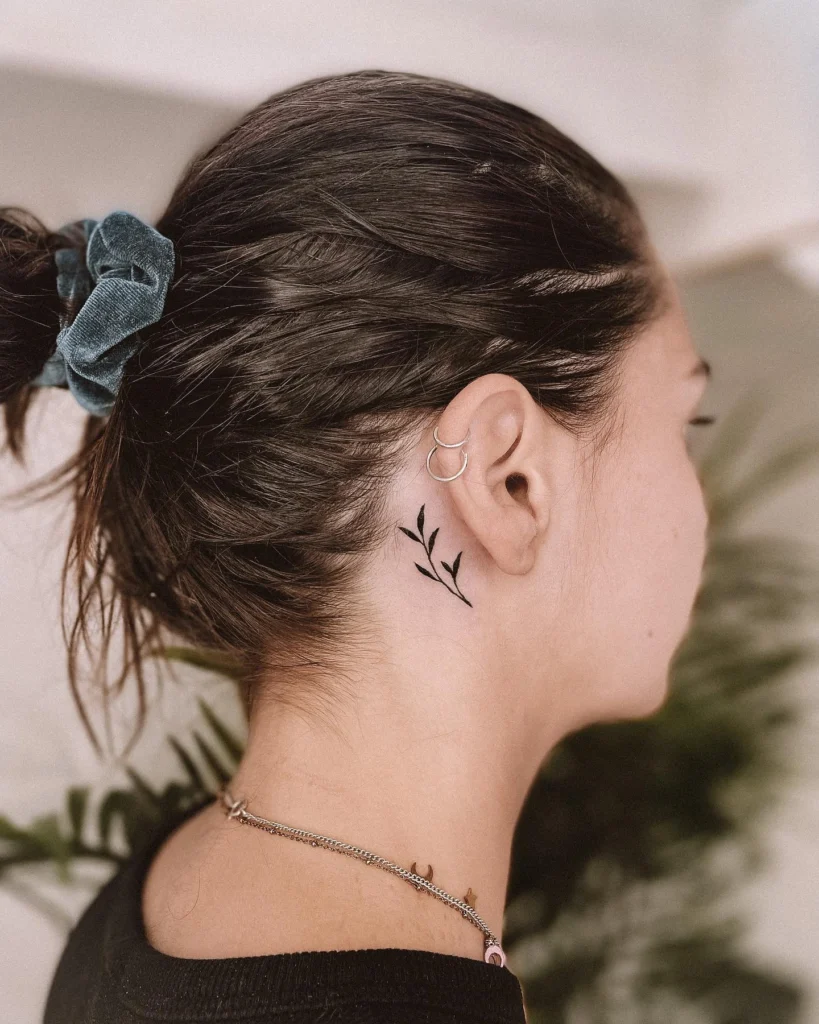 Side view of a minimalist leaf tattoo behind the ear, paired with small silver hoop earrings and a scrunchie in the hair.