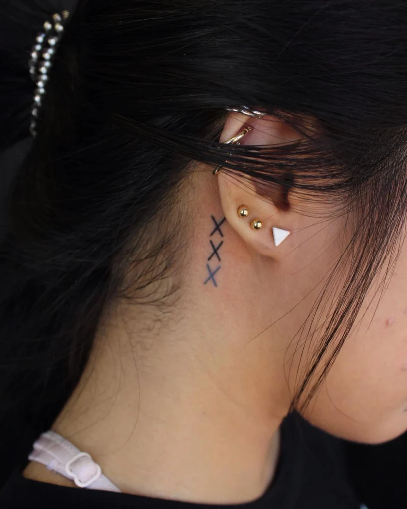 Side view of a triple "X" tattoo behind the ear, paired with gold and geometric ear piercings.