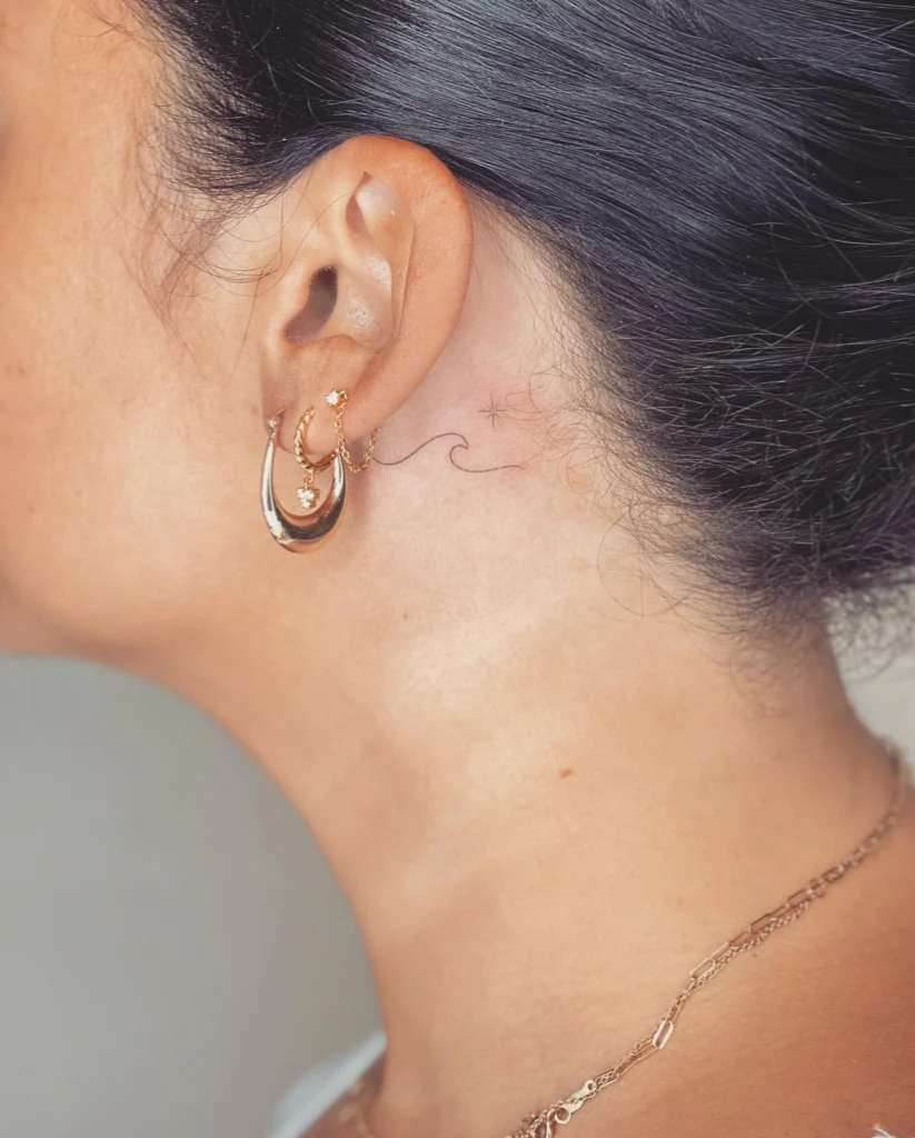 Side view of a fine line wave and tiny star tattoo behind the ear, paired with gold hoop earrings.