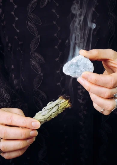 "Person using Celestite crystal in a meditation ritual, holding the stone in one hand and a smudging sage stick in the other to cleanse the crystal’s energy.