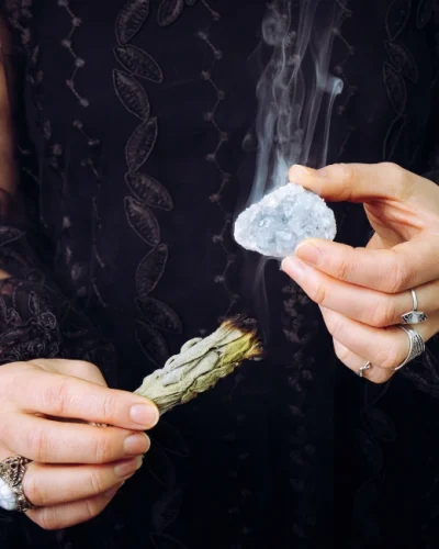 "Person using Celestite crystal in a meditation ritual, holding the stone in one hand and a smudging sage stick in the other to cleanse the crystal’s energy.