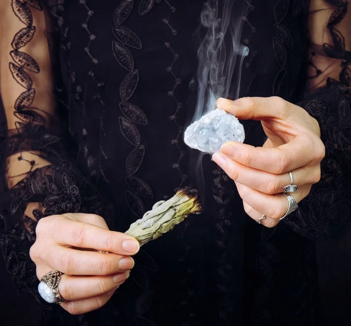 "Person using Celestite crystal in a meditation ritual, holding the stone in one hand and a smudging sage stick in the other to cleanse the crystal’s energy.