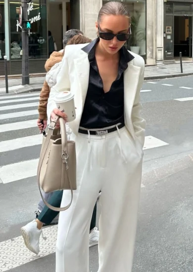 A woman wearing a white suit with a black shirt, sunglasses, and carrying a beige handbag, showcasing a sophisticated and elegant look that epitomizes Classic Style Clothing.