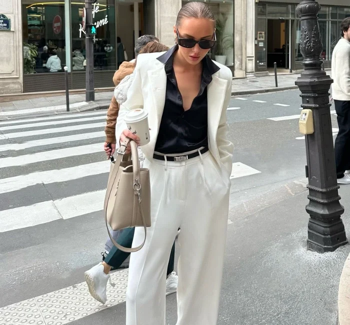 A woman wearing a white suit with a black shirt, sunglasses, and carrying a beige handbag, showcasing a sophisticated and elegant look that epitomizes Classic Style Clothing.