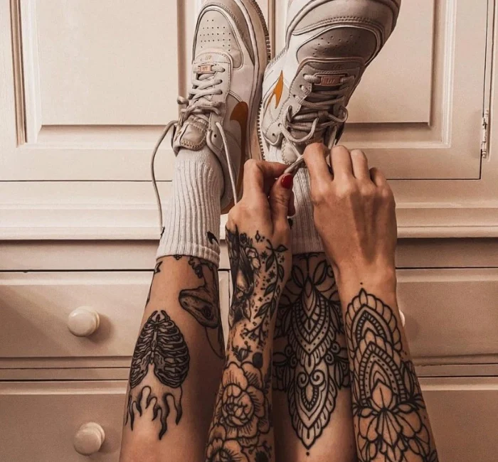 Person with intricate shin tattoos tying the laces of white sneakers, featuring a mix of mandala and anatomical designs on both legs.