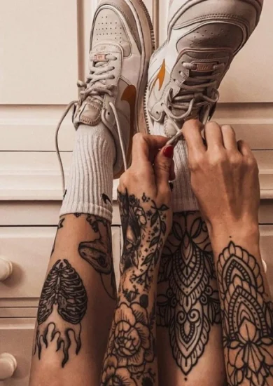 Person with intricate shin tattoos tying the laces of white sneakers, featuring a mix of mandala and anatomical designs on both legs.