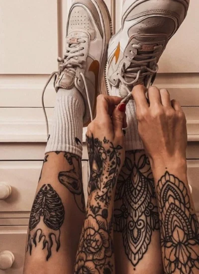 Person with intricate shin tattoos tying the laces of white sneakers, featuring a mix of mandala and anatomical designs on both legs.