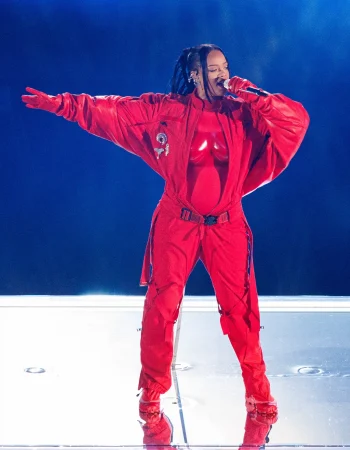 Rihanna dressed in a striking, all-red ensemble, featuring a bold oversized jacket, gloves, and cargo pants, as they sing on stage. The outfit exudes a powerful and confident energy, with the performer commanding the stage with their dynamic presence. The bright red color and stylish design make the outfit a standout fashion statement.